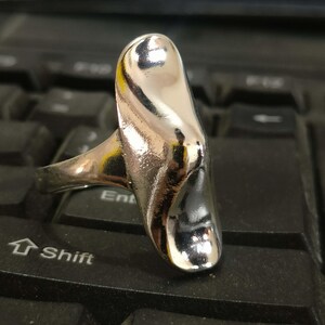 May include: A silver-toned ring featuring a smooth, elongated, and vertically oriented metal centerpiece with a polished, reflective surface. The design consists of a curved, organic shape that tapers slightly toward the top and bottom edges.