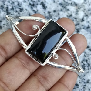 May include: A silver cuff bracelet featuring a large, rectangular, polished black stone set in the center. The metal band is crafted with open, curved wirework details that extend from the central setting to the sides of the bracelet.