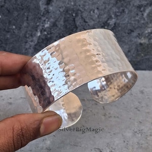 May include: A wide, silver cuff bracelet with a hammered texture. The open-ended design allows for easy wear. The bracelet is made of a shiny, reflective metal. The text "SilverBigMagic" is visible.