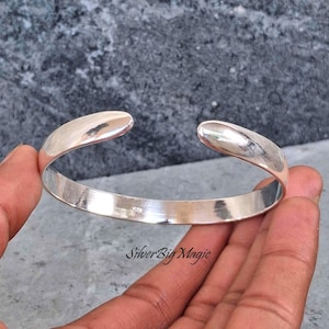 May include: A polished silver cuff bracelet with rounded ends. The open design allows for easy wear. The bracelet is held in a hand against a grey background. The text "SilverBig Magic" is visible on the bottom right.