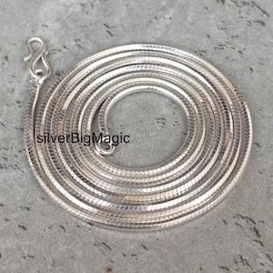 Puede incluir: Un collar de cadena de serpiente de color plateado enrollado en un patrón circular, con un acabado metálico pulido y un cierre de gancho en S. El texto silverBigMagic es visible en el lado izquierdo de la cadena de eslabones lisos y flexibles.
