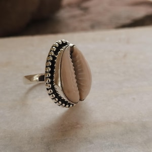 May include: A silver-toned ring featuring a central, smooth, light beige cowrie shell. The shell is framed by a decorative, beaded metal border that encircles the oval setting, attached to a plain, polished silver-toned metal band.