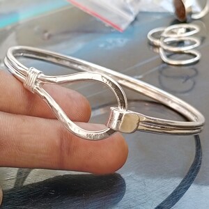 May include: A polished silver-tone metal bangle bracelet featuring a teardrop-shaped loop closure at the front. The bracelet is constructed from two parallel wires that meet at a central clasp mechanism, creating a continuous, smooth, and reflective surface.