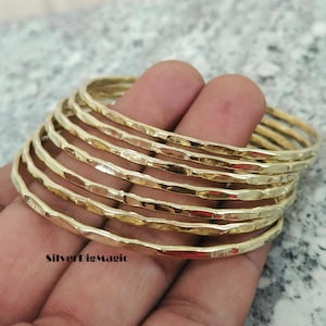 Peut inclure: Un ensemble de sept bracelets joncs en métal doré présentant une surface martelée et texturée. Chaque bracelet a un design circulaire fin avec une finition réfléchissante. Le texte SilverBigMagic est imprimé en noir dans le coin inférieur gauche.