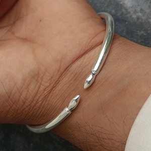 May include: A polished silver-tone open cuff bracelet featuring a smooth, rounded band that tapers into two distinct, pointed, teardrop-shaped terminals at the opening. The metal has a reflective, metallic finish and a simple, clean design.