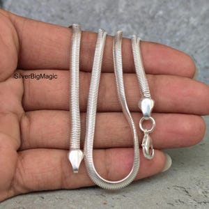 Op de afbeelding: Een zilveren slangenketting met een glad, flexibel en strak geschakeld metalen ontwerp. De ketting is voorzien van een gepolijste zilveren karabijnsluiting en een plat, taps toelopend uiteinde. De tekst SilverBigMagic is links zichtbaar.