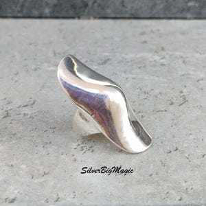 May include: A polished silver ring with a flowing, organic design. The ring has a wide, curved top that tapers to a point. The ring is set against a gray background. The text "SilverBigMagic" is visible at the bottom.