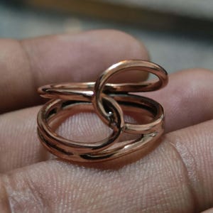 May include: A polished copper-coloured ring featuring a continuous, overlapping design that creates two distinct circular loops. The metal surface is smooth and reflective, showcasing a warm metallic tone throughout the entire band structure.
