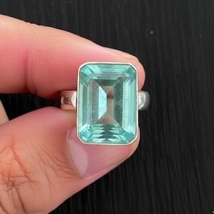 Peut inclure: Bague rectangulaire en pierre précieuse bleu clair avec un anneau argenté. La pierre précieuse est de couleur aigue-marine claire et la bague est tenue par une main. La bague est une pièce de déclaration, parfaite pour ajouter une touche de couleur.