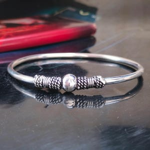Może przedstawiać: Srebrzysta bransoletka typu bangle z gładką, polerowaną centralną kulą, otoczoną dwiema sekcjami o skomplikowanych, skręconych metalowych detalach przypominających linę. Bransoletka ma cienki, okrągły kształt z odblaskowym, metalicznym wykończeniem.