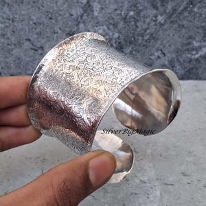 May include: A wide, silver cuff bracelet with an intricate floral pattern. The open design allows for easy wear. The bracelet is polished and reflects light, showcasing the detailed design. The text "SilverBigMagic" is visible.