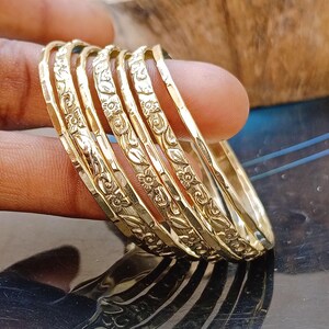 May include: A set of five gold-toned metal bangles featuring a repeating embossed floral and leaf pattern. Each bangle has a polished, reflective surface with a consistent width and a rounded profile, creating a stacked appearance on the wrist.