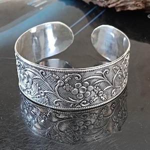 May include: A wide silver cuff bracelet featuring an intricate embossed floral pattern with vines and leaves. The metal has a polished finish on the interior and a textured, oxidized appearance on the exterior surface, showcasing detailed botanical motifs.
