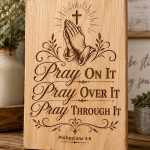 May include: A rectangular light brown wooden plaque featuring a laser-engraved design of praying hands beneath a cross. The text reads: Pray On It, Pray Over It, Pray Through It, and Philippians 4:6, surrounded by decorative scrollwork.