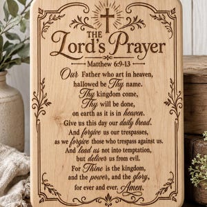 May include: A rectangular light-coloured wooden plaque featuring a laser-engraved design of The Lord's Prayer. The text is centred with a cross at the top and decorative floral scrollwork borders. The wood grain is visible throughout the surface.