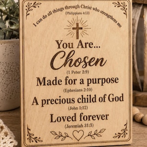 Op de afbeelding: Een rechthoekig, lichtgekleurd houten bord met lasergegraveerde tekst: I can do all things through Christ who strengthens me (Philippians 4:13), You Are... Chosen (1 Peter 2:9), Made for a purpose (Ephesians 2:10), A precious child of God (John 1:12), Loved forever (Jeremiah 31:3), inclusief een kruis- en hartsymbool.