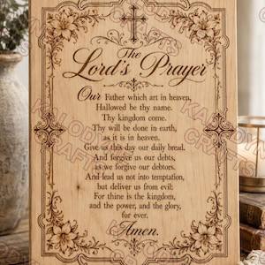 May include: A rectangular light-coloured wooden plaque featuring a laser-engraved Lord's Prayer. The text is centred within an ornate border decorated with floral motifs and crosses at the top and sides. The wood grain is visible throughout the piece.