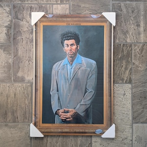 May include: A framed portrait of a man in a light gray suit and blue shirt. The portrait is labeled "THE KRAMER" at the top and includes a quote at the bottom. The frame is a light brown color.