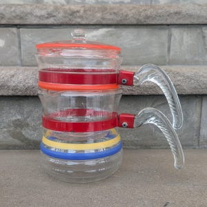 May include: A clear glass teapot with a red lid and red bands. The teapot has a clear glass handle and is decorated with yellow and blue bands. The teapot is designed to be stacked.