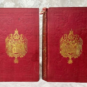 K&ouml;nnte beinhalten: Zwei antike B&uuml;cher mit roten Einb&auml;nden und goldgepr&auml;gten Designs. Die Einb&auml;nde weisen ein detailliertes Wappen und dekorative Schn&ouml;rkel auf. Die Buchr&uuml;cken sind sichtbar und zeigen den Titel und Abnutzungserscheinungen. Die B&uuml;cher stammen wahrscheinlich aus dem sp&auml;ten 19. oder fr&uuml;hen 20. Jahrhundert.