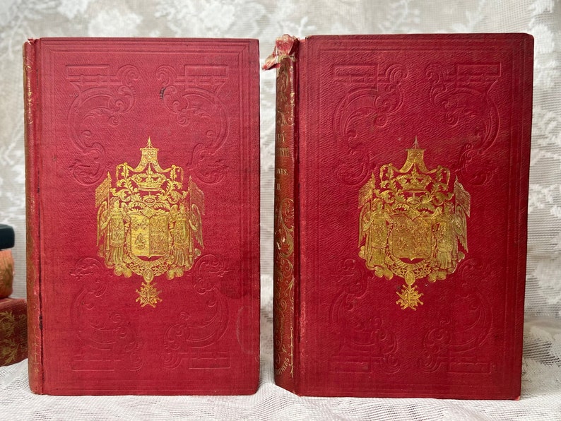 K&ouml;nnte beinhalten: Zwei antike B&uuml;cher mit roten Einb&auml;nden und goldgepr&auml;gten Designs. Die Einb&auml;nde weisen ein detailliertes Wappen und dekorative Schn&ouml;rkel auf. Die Buchr&uuml;cken sind sichtbar und zeigen den Titel und Abnutzungserscheinungen. Die B&uuml;cher stammen wahrscheinlich aus dem sp&auml;ten 19. oder fr&uuml;hen 20. Jahrhundert.