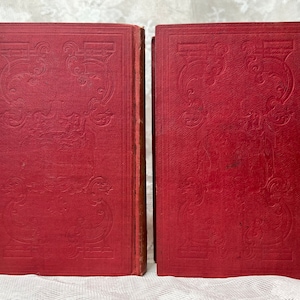K&ouml;nnte beinhalten: Zwei antike B&uuml;cher mit roten Einb&auml;nden. Die Einb&auml;nde weisen ein gepr&auml;gtes dekoratives Design auf. Die B&uuml;cher stehen nebeneinander aufrecht, wobei ihre Buchr&uuml;cken nach au&szlig;en zeigen. Die Kanten der Seiten sind sichtbar.