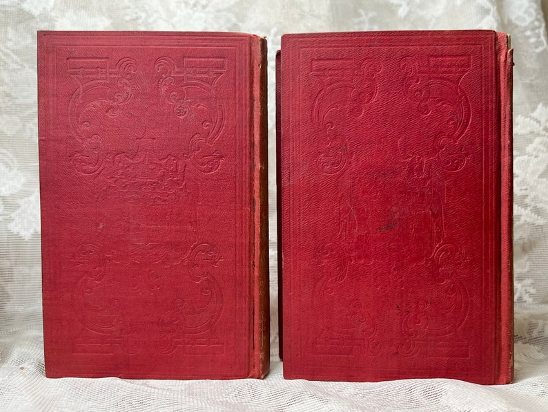 K&ouml;nnte beinhalten: Zwei antike B&uuml;cher mit roten Einb&auml;nden. Die Einb&auml;nde weisen ein gepr&auml;gtes dekoratives Design auf. Die B&uuml;cher stehen nebeneinander aufrecht, wobei ihre Buchr&uuml;cken nach au&szlig;en zeigen. Die Kanten der Seiten sind sichtbar.