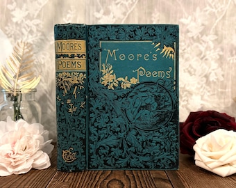 Könnte beinhalten: Ein tealfarbenes Hardcover-Buch mit dem Titel Moore's Poems, verziert mit schwarzen und goldenen floralen Prägemustern sowie einer kreisförmigen Schwanenillustration auf dem Einband. Der Buchrücken zeigt goldene Schrift und florale Motive.