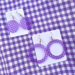 Peut inclure: Deux paires de boucles d'oreilles sur des cartons blancs, sur un fond à carreaux vichy violets et blancs. Une paire présente des boucles d'oreilles rondes et pleines avec un motif vichy violet. L'autre paire a de grandes boucles d'oreilles créoles avec le même motif vichy.