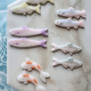 May include: A collection of decorative fish figurines in various colors. The fish include yellow, pink, and white with orange spots. The figurines are arranged on a light-colored surface, with a water-like background.