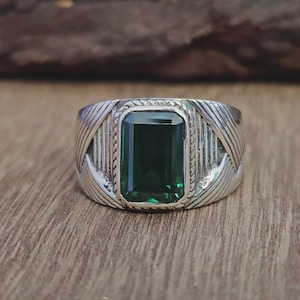 May include: A silver ring featuring a rectangular, deep green emerald. The ring has a textured band with a woven pattern. The emerald is set in a silver bezel, showcasing its vibrant colour.