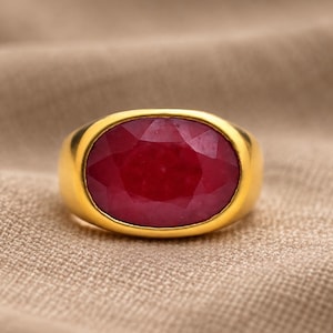 Puede incluir: Un anillo de oro con una gran piedra preciosa de color rojo intenso, forma ovalada y tallada en facetas, engastada en una banda de oro lisa y pulida. La piedra muestra un tono carmesí rico y saturado con facetas internas que captan la luz.