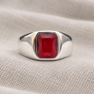 May include: A silver-toned ring featuring a large, rectangular, faceted red gemstone set in the center. The band is smooth and polished, with a wide, solid construction that tapers slightly toward the back, highlighting the vibrant red stone.