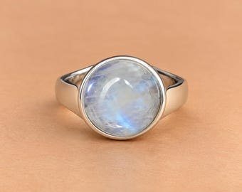 Puede incluir: Un anillo de plata con una piedra lunar redonda de corte cabujón engastada en un bisel liso. La gema presenta un aspecto blanco lechoso y translúcido con una sutil iridiscencia azul, y la banda de plata pulida tiene un diseño sencillo.