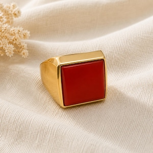 May include: A gold-toned ring featuring a large, flat, square-shaped red stone set into a polished metal band. The ring has a thick, solid construction with a smooth, reflective surface and a prominent, vibrant red center piece.
