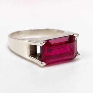 May include: A silver-toned ring featuring a large, rectangular, emerald-cut vibrant red gemstone set horizontally. The metal band is smooth and polished, with four prongs securing the faceted red stone in a clean, open-sided setting design.