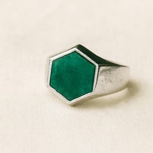 Puede incluir: Anillo de plata con una piedra hexagonal verde esmeralda. El anillo tiene un diseño sencillo y moderno con un acabado pulido. La piedra preciosa está engastada en un bisel de plata, creando un contraste llamativo. Una joya elegante.