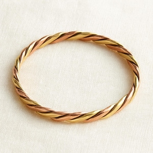 Pode incluir: Uma pulseira de metal torcido com um design trançado. A pulseira apresenta seções alternadas de metal dourado e cor de cobre, criando um efeito de dois tons. A pulseira é circular e parece ser feita de um material durável.