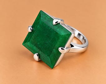 May include: A silver-toned ring featuring a large, square-cut, deep green gemstone set with four prominent prongs. The band is smooth and polished, extending from the base of the square setting to form a continuous, reflective metallic loop.