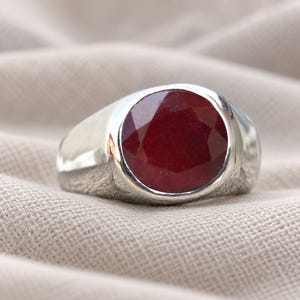Könnte beinhalten: Ein silberfarbener Ring mit einem großen, runden, facettierten tiefroten Edelstein, der in eine glatte, polierte Metallschiene gefasst ist. Der Edelstein zeigt einen dunklen Karmesinton mit sichtbaren Facetten, während die Schiene massiv ist.