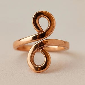 May include: A copper-colored ring with a unique, swirling design. The ring features two rounded loops connected by a band that wraps around the finger. The metal has a polished, reflective surface, creating a warm, inviting aesthetic. This piece is a stylish accessory.