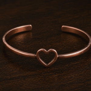 May include: A copper-colored cuff bracelet with an open heart design in the center. The bracelet has a smooth, polished finish and a simple, elegant style. The heart is a cutout shape, adding a delicate touch to the jewelry.