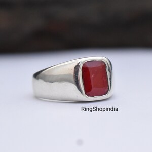 May include: A silver ring featuring a red gemstone. The ring has a plain design and is set with a square-shaped stone.