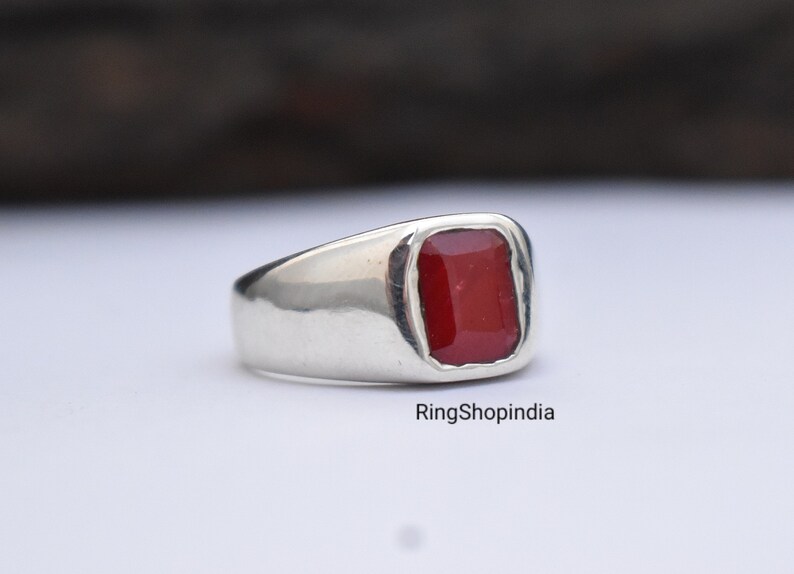 May include: A silver ring featuring a red gemstone. The ring has a plain design and is set with a square-shaped stone.