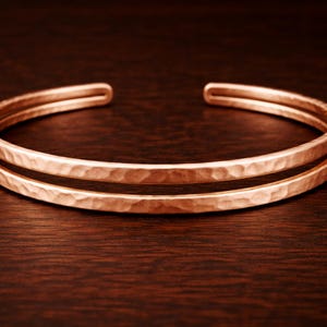 May include: A copper cuff bracelet with a hammered texture. The open-ended bracelet features two parallel bands. The warm, reddish-brown metal reflects light, creating highlights and shadows. The bracelet is displayed on a dark wood surface.