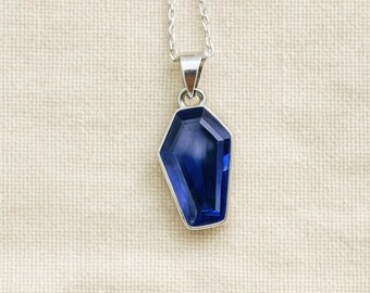 May include: A silver necklace with a pendant featuring a deep blue, coffin-shaped gemstone. The gemstone is set in a silver bezel and hangs from a delicate silver chain. The background is a neutral, textured fabric.