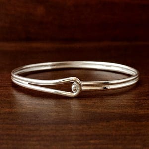 Op de afbeelding: Een zilverkleurige armband met een haak- en oogsluiting. De armband heeft een eenvoudig, elegant ontwerp met een gladde, gepolijste afwerking. De haaksluiting heeft een kleine, ronde bal. De armband wordt getoond op een donkerbruin houten oppervlak.
