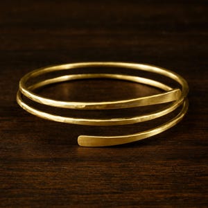 May include: A gold-colored, hammered-finish bracelet with a spiral design. The bracelet wraps around the wrist with three bands, creating a layered effect. The open end allows for easy wear. The bracelet is displayed on a dark wood surface.
