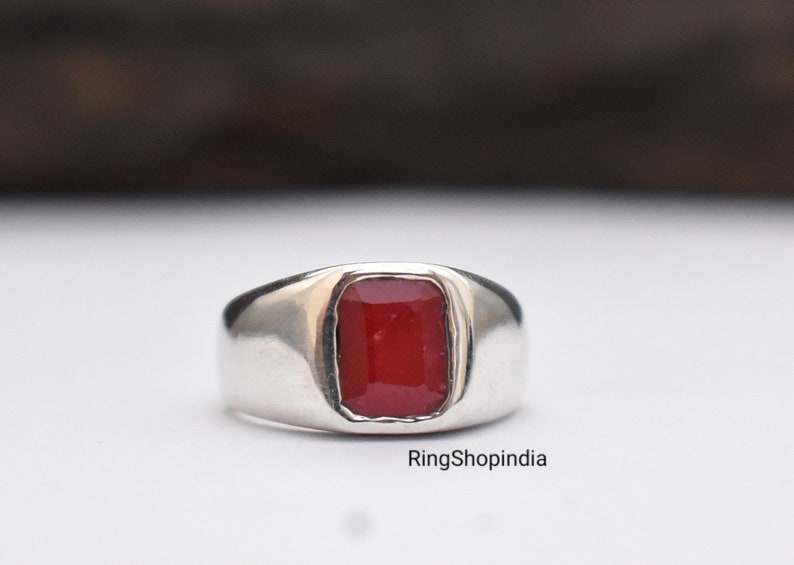 May include: A silver ring with a red gemstone set in a square shape. The ring has a wide band and a polished finish.