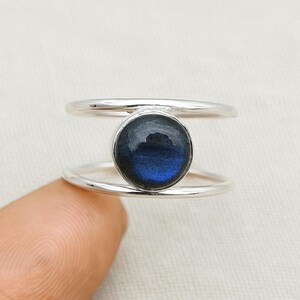 May include: A silver ring featuring a round, dark blue gemstone with iridescent qualities. The ring has a double-band design, with the gemstone set between two parallel bands. The ring is shown on a finger.